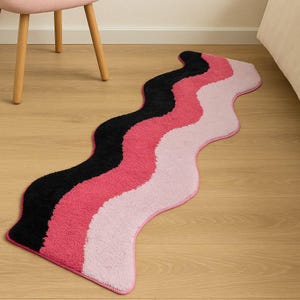 May include: A wavy rug with a black, hot pink, and light pink color scheme. The rug has a pink border and is placed on a wooden floor. A pink chair and a bed are in the background.