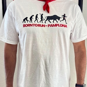 May include: White t-shirt with a black and red graphic depicting the evolution of a runner, a bull, and the text "BORN TO RUN - PAMPLONA". A red scarf is tied around the neck.