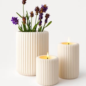 Könnte beinhalten: Drei cremefarbene, gerippte Keramikobjekte auf weißem Hintergrund. Eine hohe Vase enthält lila Lavendel. Zwei zylindrische Kerzen brennen und erzeugen ein warmes Leuchten. Die Anordnung erzeugt eine minimalistische Ästhetik.