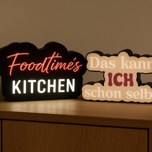 Könnte beinhalten: Zwei beleuchtete Schilder mit schwarzen Rahmen. Eines lautet "Foodtime's KITCHEN" in Rot und Weiß. Das andere sagt "Das kann ICH schon selber" in Weiß und Burgunder. Die Schilder stehen auf einer Holzoberfläche.