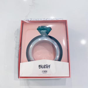 May include: A silver bracelet flask shaped like a diamond ring, with a teal-colored gem on top. The flask is in a pink box with the text "BLUSH I DO! and bracelet flask."