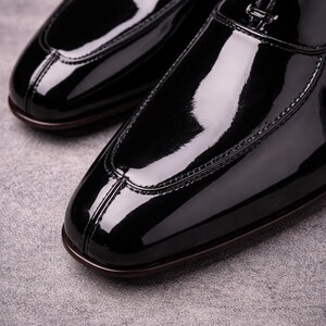 May include: Close-up of two black patent leather dress shoes. The shoes have a glossy finish, a rounded toe, and a stitched design along the edges. The soles are a dark brown colour. The shoes are on a grey surface.