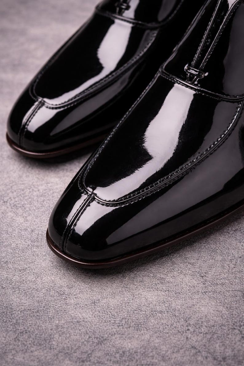 May include: Close-up of two black patent leather dress shoes. The shoes have a glossy finish, a rounded toe, and a stitched design along the edges. The soles are a dark brown colour. The shoes are on a grey surface.