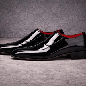 May include: A pair of black patent leather dress shoes with a glossy finish. The shoes have a low heel and a red interior lining. The shoes are displayed on a grey surface.