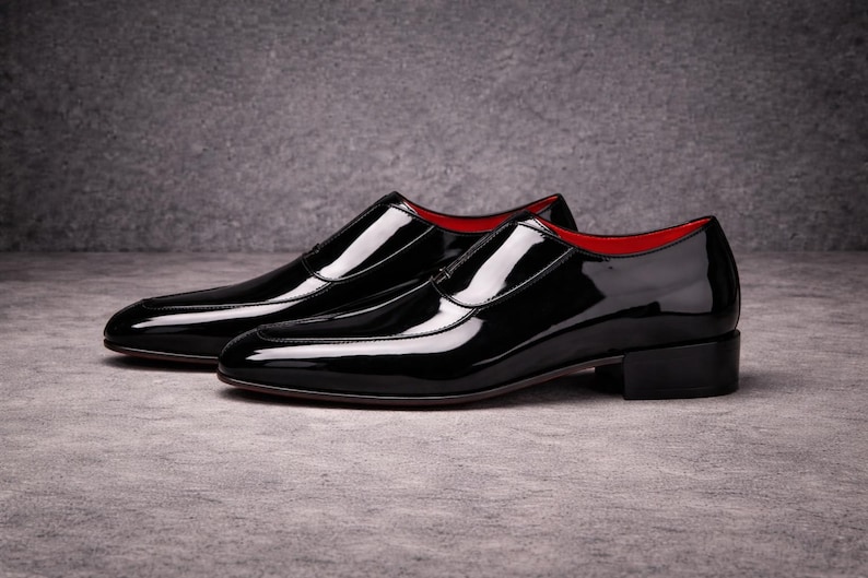May include: A pair of black patent leather dress shoes with a glossy finish. The shoes have a low heel and a red interior lining. The shoes are displayed on a grey surface.