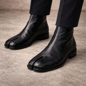 May include: Black leather ankle boots with a split-toe design. The boots have a sleek, minimalist aesthetic, featuring a low heel and a smooth, polished finish. The boots are paired with black trousers.