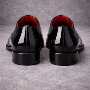 May include: Two black patent leather dress shoes with a high-gloss finish. The shoes feature a classic design with a low heel and a red interior lining. The image is shot from the rear, highlighting the sleek silhouette.