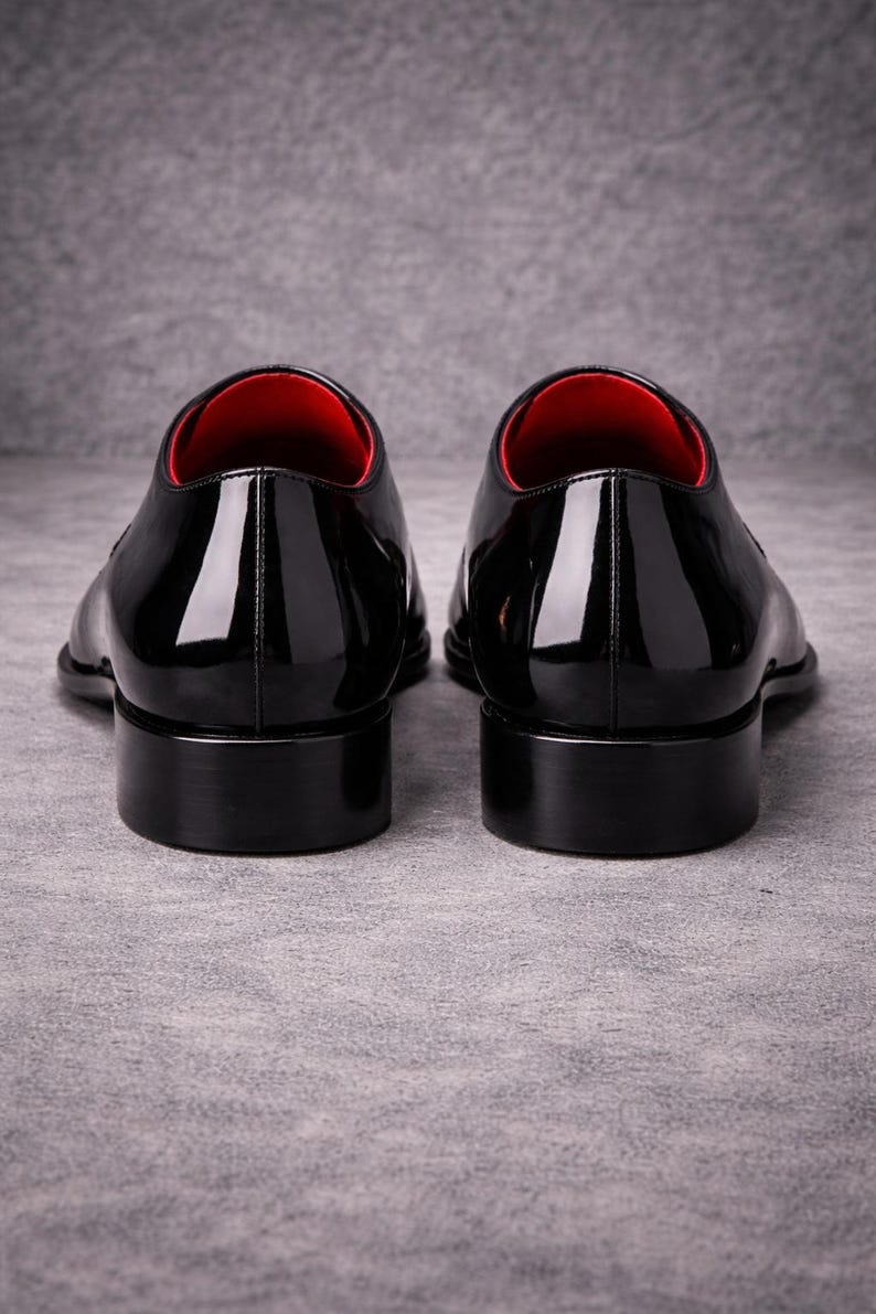 May include: Two black patent leather dress shoes with a high-gloss finish. The shoes feature a classic design with a low heel and a red interior lining. The image is shot from the rear, highlighting the sleek silhouette.