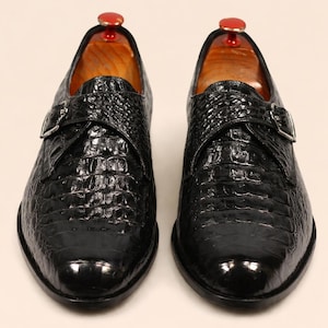 Könnte beinhalten: Paar schwarze Lederschuhe mit Krokodilleder-Textur. Die Schuhe haben einen Schnallenverschluss und werden mit Schuhspannern aus Holz präsentiert. Klassischer Stil.