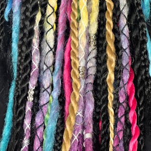 Può includere: Primo piano di dreadlocks colorati in varie tonalità di blu, nero, giallo, rosa, viola e oro. Alcuni dreadlocks sono adornati con perline di metallo argentato e avvolgimenti di filo, creando un aspetto strutturato e vibrante. I dreadlocks sono attorcigliati.