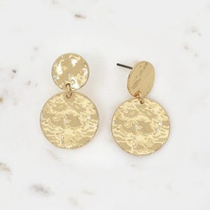 以下が含まれることがあります： ハンマー加工されたテクスチャのゴールドカラーのイヤリングのペア。各イヤリングは、小さな丸いスタッドと、より大きな円形のペンダントで構成されています。イヤリングは白い大理石の表面に表示されています。
