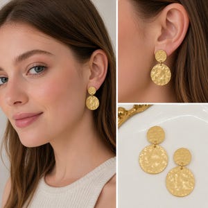 Può includere: Orecchini color oro con texture martellata. Ogni orecchino presenta un piccolo perno rotondo collegato a un pendente circolare più grande. Gli orecchini sono mostrati sull'orecchio di una persona e anche separatamente su una superficie bianca.