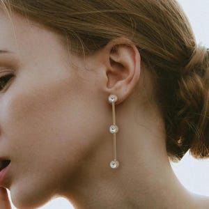 May include: Gold-toned drop earrings with three clear, round crystals. The earrings hang vertically, with each crystal set in a bezel. The design is simple and elegant, suitable for various occasions.
