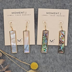 Może przedstawiać: Dwie pary prostokątnych kolczyków wiszących. Jedna para ma perłową białą wkładkę, druga kolorowy wzór muszli abalonowej. Oba mają złote ramki i haczyki na uszy. Kolczyki są prezentowane na białych kartach z napisem "MOMENT.J JEWEL YOUR MOMENT".