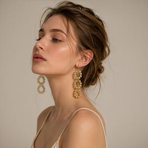May include: A pair of gold-toned drop earrings featuring a vertical arrangement of three interconnected circular links. Each link is constructed from a series of small, spherical beads, creating a textured, metallic surface that catches the light.