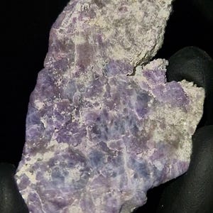 May include: A raw, irregular mineral specimen featuring a mix of vibrant purple and soft lavender crystalline sections embedded within a rough, pale grey, and white rocky matrix. The stone has a jagged, uneven surface texture with visible mineral deposits.