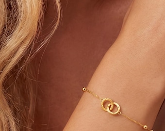 May include: A delicate gold chain bracelet featuring two interlocking circular rings as a central focal point, accented by small gold beads spaced evenly along the thin chain, secured with a clasp, creating a refined and polished jewelry piece.