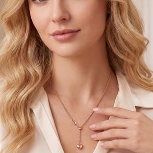 May include: A rose gold-toned necklace featuring a delicate chain and a small, smooth heart-shaped pendant. The necklace includes an adjustable sliding bead mechanism that allows the chain to hang in a Y-shape, resting against a cream-colored blouse.
