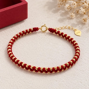 Op de afbeelding: Een rood koordarmbandje met een reeks kleine, ronde, goudkleurige kralen die in het koord zijn geweven. Het armbandje is voorzien van een ronde, goudkleurige veerringsluiting, een korte verlengketting en een klein hartvormig label met de inscriptie AU750.