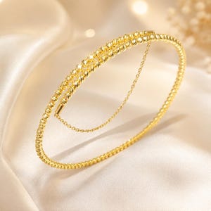 Puede incluir: Una pulsera rígida de oro con un diseño de cuentas facetadas y texturizadas que crea un efecto brillante. La pulsera incluye una delicada y fina cadena de oro que conecta dos puntos de la banda circular, añadiendo un aspecto superpuesto a la pieza.