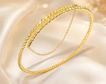 May include: A gold bangle bracelet featuring a textured, faceted bead design that creates a shimmering effect. The bracelet includes a delicate, thin gold chain that connects two points of the circular band, adding a layered appearance to the piece.