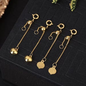 May include: Four gold-toned jewelry extenders, each featuring a spring ring clasp, a sliding bead, a delicate chain, and a heart-shaped charm at the end. The charms on the right two extenders display a small engraved stamp on their surface.