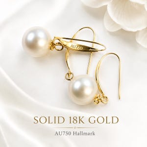Può includere: Un paio di orecchini pendenti con perle bianche rotonde sospese a ganci in oro giallo. I ganci in oro riportano la scritta AU750. Il testo SOLID 18K GOLD e AU750 Hallmark è stampato sotto gli orecchini sulla superficie in tessuto.