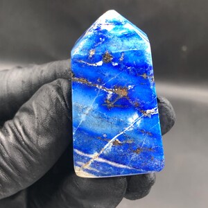 May include: A polished lapis lazuli obelisk, rectangular in shape. The stone is a deep blue with white and gold veins. The top is a lighter blue. The obelisk is held by a gloved hand against a dark background.