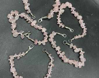 May include: Multiple necklaces and bracelets crafted from pink rose quartz chips, strung on white cord. Each piece features a silver-coloured clasp. The jewellery is displayed on a dark surface.