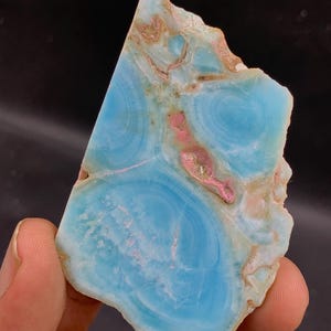 May include: A polished, irregularly shaped gemstone with a striking blue hue, reminiscent of the ocean. The stone features swirling patterns in shades of pink, tan, and white, creating a unique and natural aesthetic. The gemstone is held in a hand against a dark background.