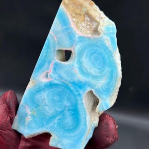 May include: A polished, triangular-shaped gemstone with a striking blue hue, featuring concentric circular patterns. The stone has a natural, rough edge with tan and pink accents, and a few natural holes. The gemstone is held by a person wearing a red glove.