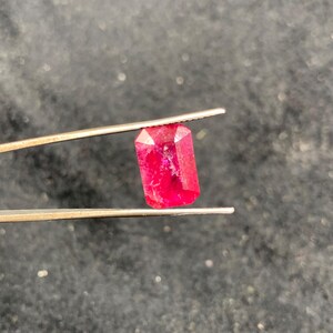 Puede incluir: Una gema de rubí rectangular y facetada sostenida por unas pinzas. El rubí es de color rojo vibrante con inclusiones internas visibles. Las pinzas son de color plateado y el fondo es una superficie oscura y borrosa.