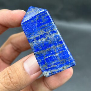 May include: A rectangular, polished lapis lazuli crystal. The stone is a deep blue colour with streaks of grey and gold. The crystal is held in a hand, showcasing its smooth, faceted surface and natural variations. The crystal is a gemstone.