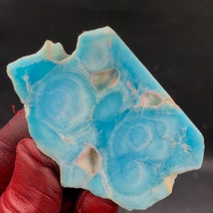 Pode incluir: Uma gema vibrante, de forma irregular, com um tom azul marcante, que lembra as águas do oceano. A pedra apresenta padrões únicos em espiral e áreas de azul mais claro e bege, criando uma aparência natural e artística. A gema é segurada por uma mão vermelha.