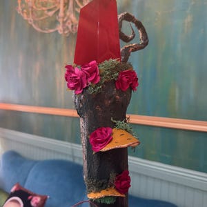 May include: A decorative wooden sculpture featuring a large, red crystal at the top. The dark brown wood is adorned with pink roses, green moss, and yellow-orange mushroom accents. A whimsical, artistic piece.
