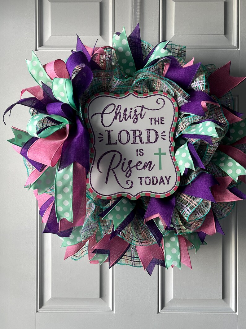 May include: An Easter wreath with a sign saying "Christ the Lord is Risen Today." The wreath is made of purple, pink, and mint green ribbons and mesh. A decorative item for the Easter holiday.
