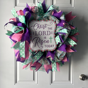 May include: An Easter wreath with a sign reading "Christ the Lord is Risen Today." The wreath is made with purple, pink, and mint green ribbons, and a mesh base. A decorative item for a front door, with a religious theme.