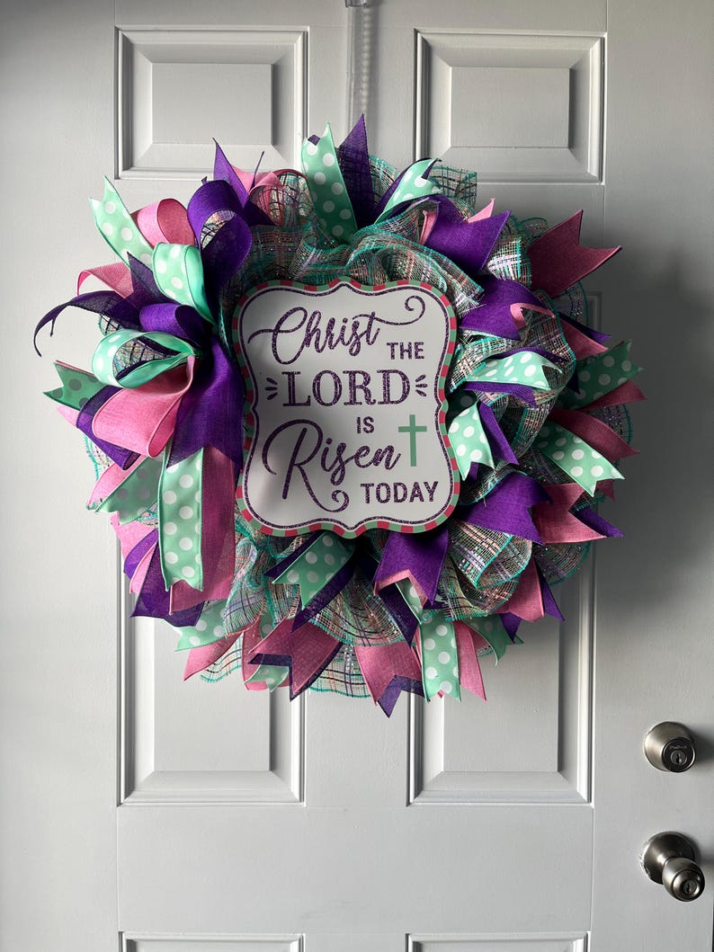 May include: An Easter wreath with a sign reading "Christ the Lord is Risen Today." The wreath is made with purple, pink, and mint green ribbons, and a mesh base. A decorative item for a front door, with a religious theme.