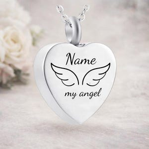May include: A silver heart-shaped pendant with the words "Name my angel" and a pair of angel wings. The pendant hangs from a delicate silver chain. The background is a soft, blurred image of white flowers.