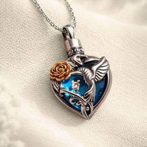 Peut inclure: Un collier pendentif en forme de cœur argenté avec une pierre centrale bleue. Le pendentif présente une rose dorée et un motif de colibri. Le collier est présenté sur une surface blanche.