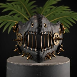 Puede incluir: Una máscara de cuero negro con un diseño steampunk. La máscara presenta púas, engranajes y costuras doradas. La máscara tiene una serie de barras verticales en la boca y está diseñada para cubrir la mitad inferior de la cara.