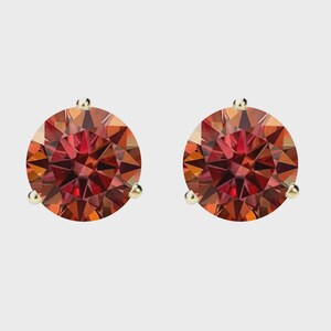May include: A pair of round, faceted, red-orange gemstone stud earrings. The gemstones are set in gold-colored prongs. The earrings are displayed against a neutral background.