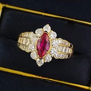May include: A gold ring featuring a marquise-cut ruby center stone, surrounded by smaller round and baguette-cut diamonds. The ring is set in a gold band and presented in a black box with a gold border. The ring's design is ornate and elegant.