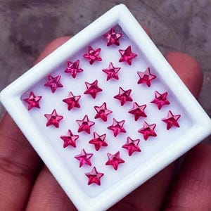 May include: A white square tray filled with small, faceted, star-shaped gemstones in a vibrant pink hue. The gemstones are arranged neatly within the tray, creating a sparkling display. The tray is held in a person's hand.