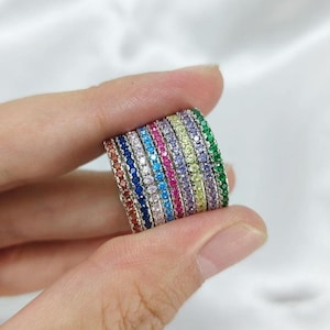May include: A collection of colorful gemstone rings. Each ring is a different color, including red, blue, pink, and green. The rings are held in a hand, showcasing the variety of colors and designs.