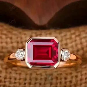 Pode incluir: Um anel de ouro com uma grande pedra preciosa retangular, vermelho escuro, no centro, ladeada por duas pedras preciosas redondas e claras menores. A banda do anel é de uma cor dourada quente, e as pedras preciosas são definidas em uma configuração de moldura.