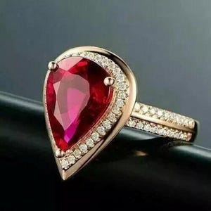 May include: Rose gold ring with a large, pear-cut, deep red gemstone. The gemstone is encircled by a halo of small, clear accent stones. The ring band also features small stones, creating a sparkling effect. A stunning piece of jewellery.