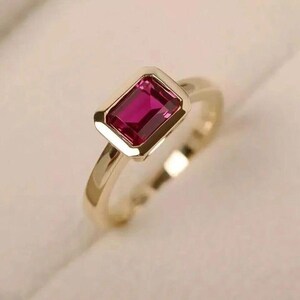 Könnte beinhalten: Ein goldener Ring mit einem rechteckigen, tiefrosa Edelstein. Der Edelstein ist in einer goldenen Fassung gefasst, und das Band ist aus poliertem Gold. Der Ring wird auf einer weichen, cremefarbenen Oberfläche gezeigt.