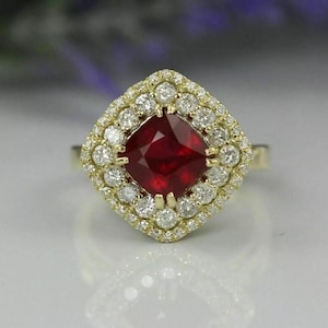 May include: A gold ring featuring a cushion-cut, deep red gemstone surrounded by a halo of smaller, round, white gemstones. The ring's setting is also gold, creating a luxurious and elegant appearance. The ring is set on a white surface.