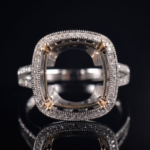 May include: A silver ring with a cushion-shaped setting, encircled by small, sparkling stones. The ring's band also features a row of smaller stones, creating a halo effect. The ring is set against a black background.